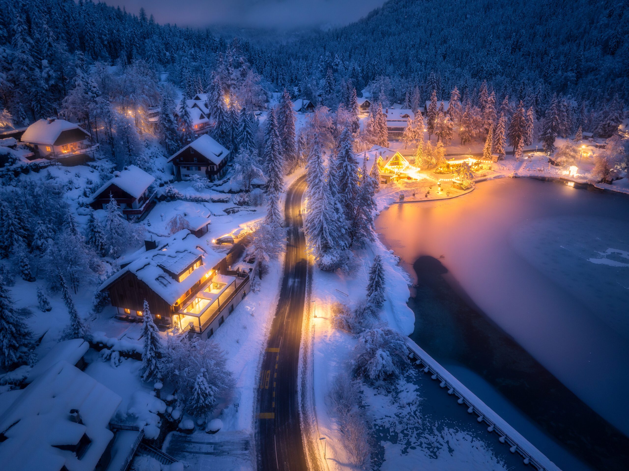 Aerial view of fairy town in snow road forest Jasna lake and houses with lights at night in winter Top view of mountain village illumination snowy pine trees at dusk in Kranjska Gora Slovenia
